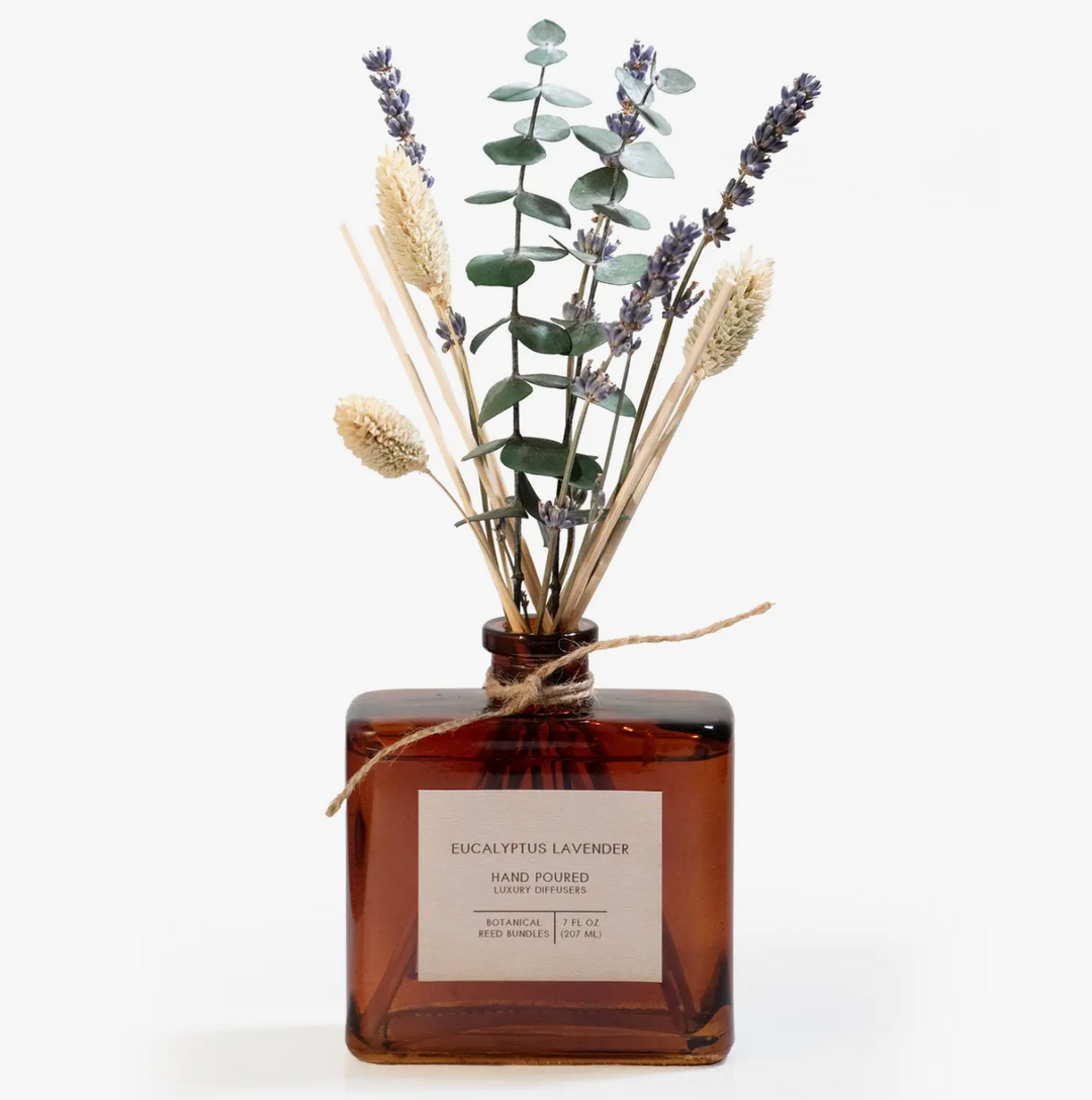 Square glass diffuser bottle filled with amber liquid, containing dried floral stems including lavender and wheat stalks, tied with twine.