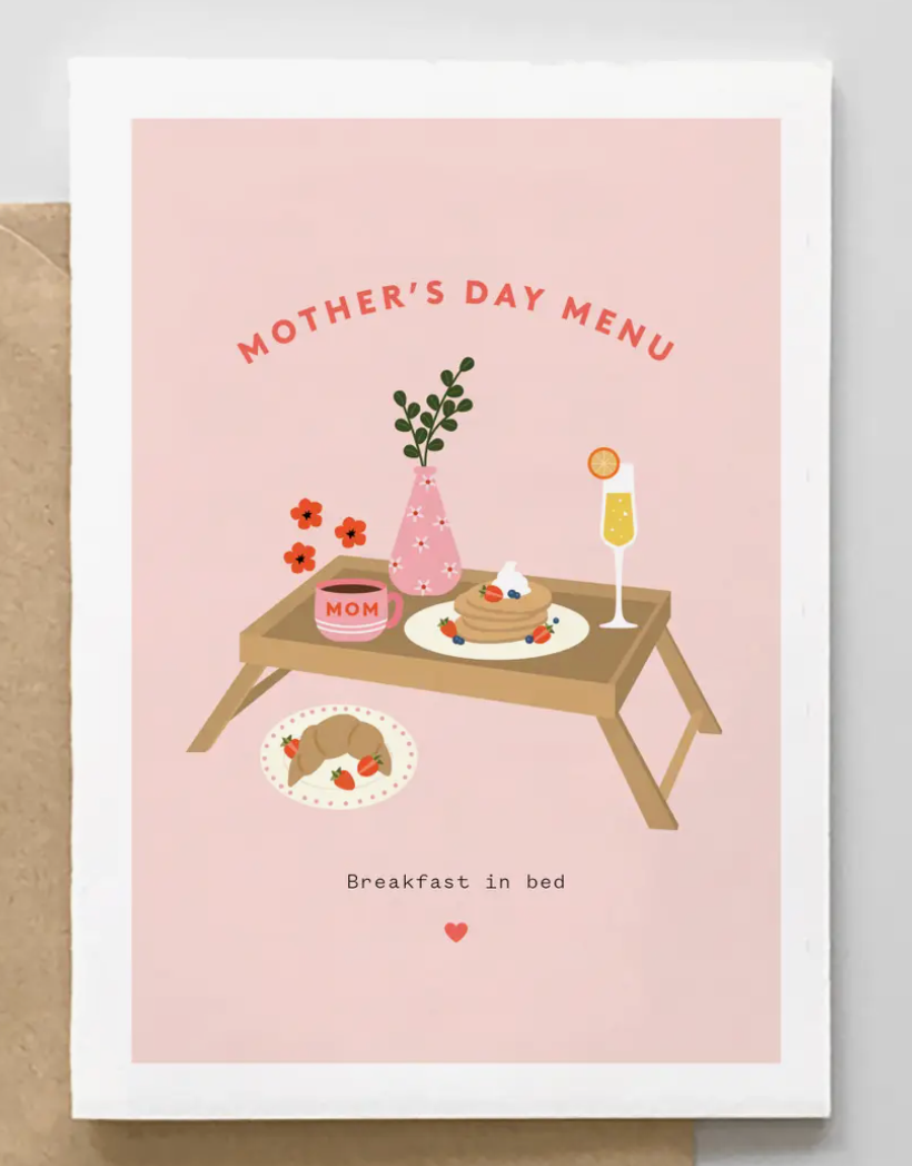 A pink greeting card with a Mother's Day menu illustration, featuring a tray with pancakes, flowers, mimosa, and a mug labeled "Mom", surrounded by a light wood frame.